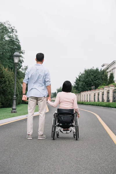 Vue arrière du jeune homme tenant la main avec une petite amie handicapée tout en marchant sur la route dans le parc — Photo de stock