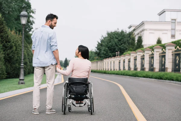 Vista trasera de hombre joven y mujer discapacitada tomados de la mano mientras caminan en el parque - foto de stock