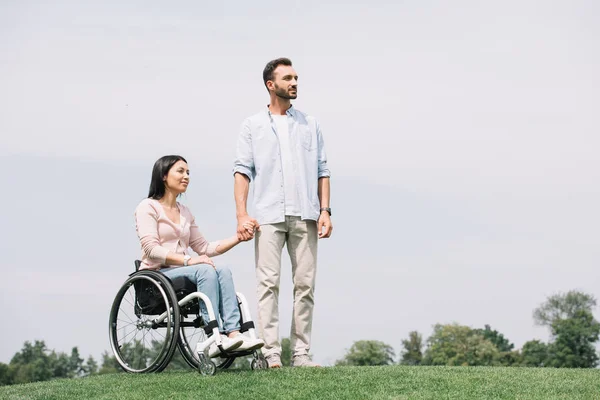 Bonito jovem homem de mãos dadas com deficiência namorada sentado em cadeira de rodas — Fotografia de Stock