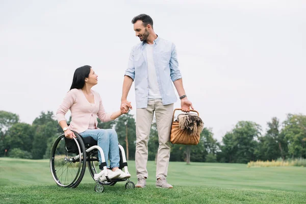 Hombre guapo con cesta de paja cogido de la mano con la novia discapacitada en el parque - foto de stock