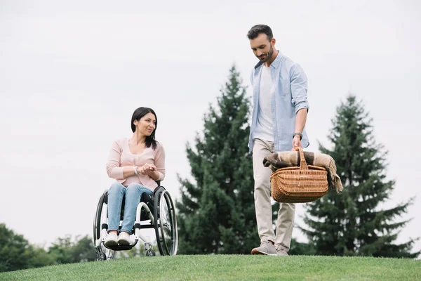 Hombre guapo sosteniendo cesta de paja cerca de novia discapacitada en el parque - foto de stock