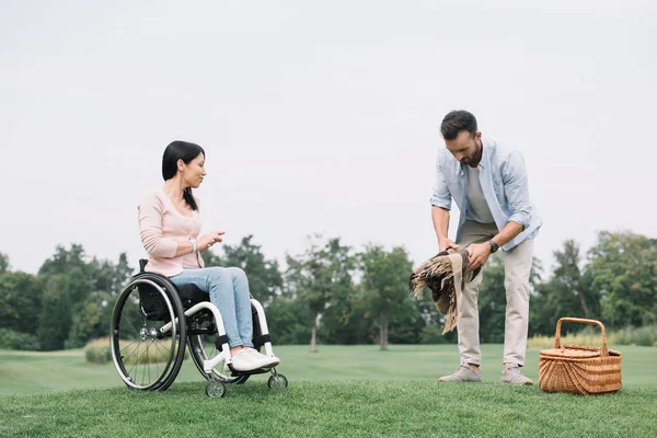 Bel homme préparant place pour pique-nique près de petite amie handicapée dans le parc — Photo de stock