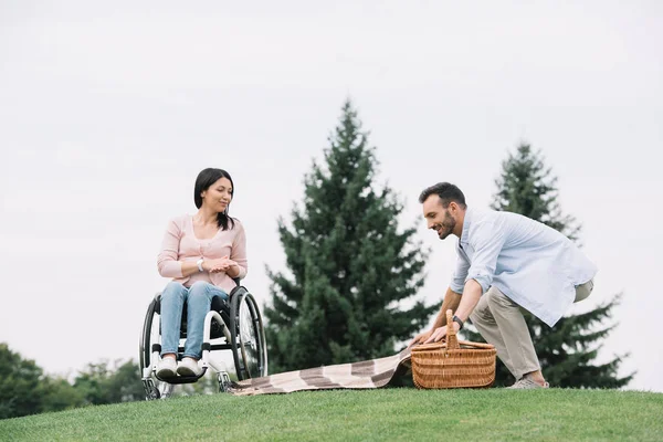 Hombre feliz lugar de preparación para el picnic cerca de novia discapacitada en el parque - foto de stock