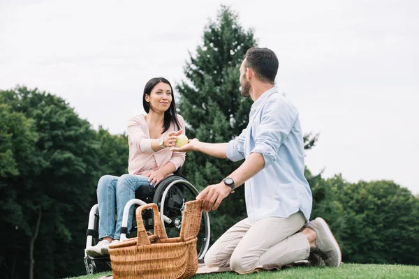 Молодой человек дарит яблоко счастливой девушке-инвалиду, отдыхая вместе в парке — стоковое фото