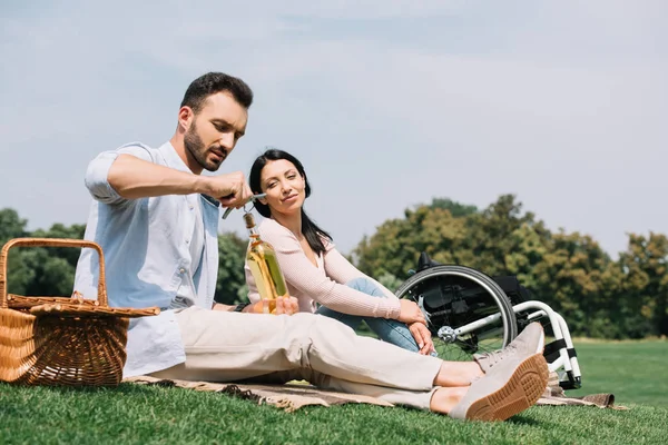 Hombre guapo apertura botella de vino blanco mientras descansa en el parque con novia discapacitada - foto de stock