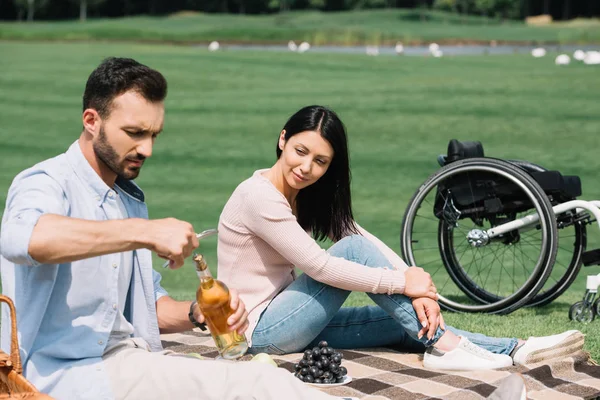 Young man opening bottle of white wine while sitting on blanket near disabled girlfriend — Stock Photo