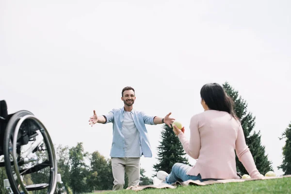 Hombre feliz gesto cerca de novia discapacitada sentado en la manta y la celebración de manzana - foto de stock