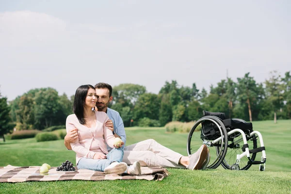 Feliz guapo hombre abrazando joven discapacitado novia mientras sentado en manta en parque - foto de stock