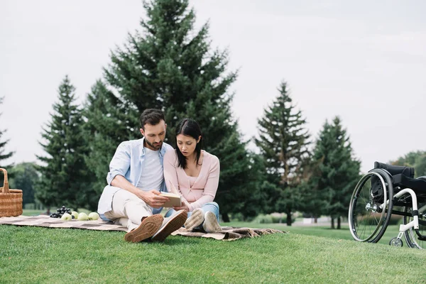 Красивый мужчина с девушкой-инвалидом читает книгу, сидя на одеяле в парке — стоковое фото