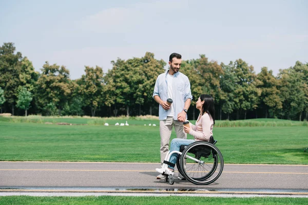 Bel homme avec petite amie handicapée tenant du café pour aller tout en marchant dans le parc — Photo de stock