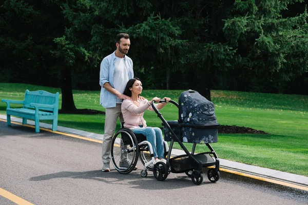 Femme heureuse en fauteuil roulant marche avec mari et bébé chariot dans le parc — Photo de stock