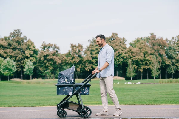 Bel homme joyeux marchant avec bébé buggy dans le parc — Photo de stock