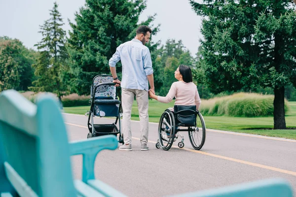 Selective focus of happy disabled woman holding hands with husband while walking with baby carriage in park together — Stock Photo