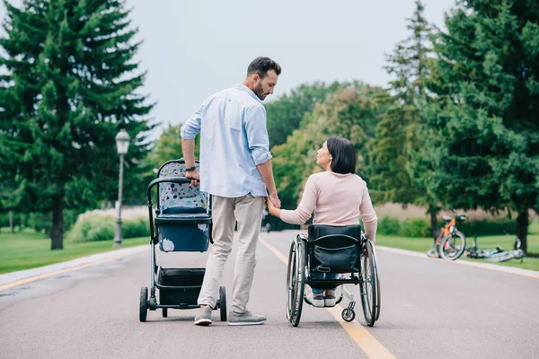 Happy man holding hand of disabled wife while walking with baby carriage in park together — Stock Photo