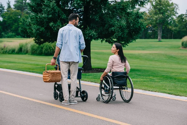 Happy disabled woman looking at husband carrying straw basket while walking in park with baby carriage together — Stock Photo