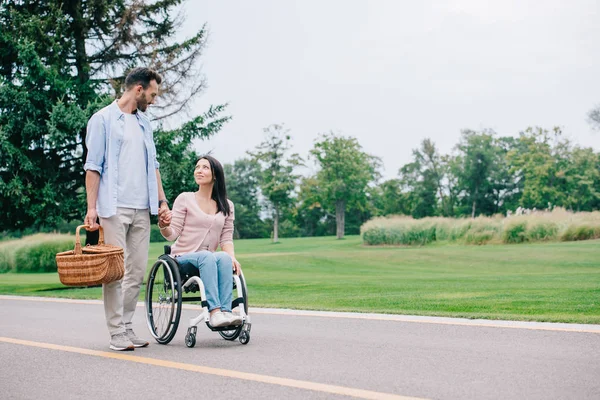 Hombre guapo con cesta de paja cogido de la mano con la novia discapacitada mientras caminan juntos en el parque - foto de stock