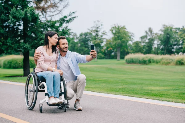 Homme heureux embrasser petite amie handicapée et prendre selfie sur smartphone dans le parc — Photo de stock