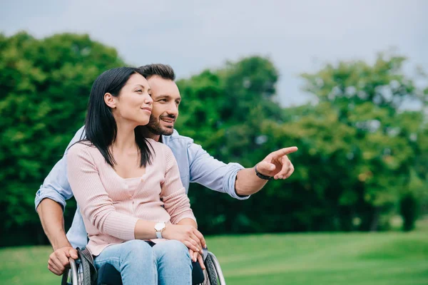 Hombre alegre señalando con el dedo mientras camina en el parque con la novia discapacitada - foto de stock