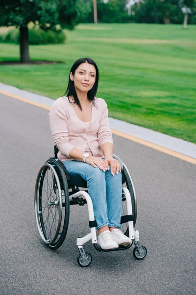 Joven mujer discapacitada en silla de ruedas sonriendo a la cámara mientras camina en el parque - foto de stock