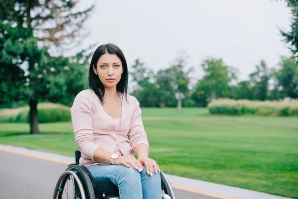 Mujer discapacitada pensativa mirando la cámara mientras está sentada en silla de ruedas en el parque - foto de stock