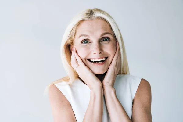 Happy blonde mature woman with hands near face isolated on grey — Stock Photo