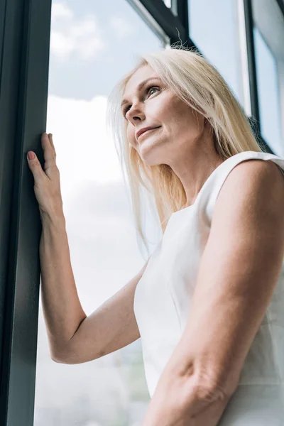 Низкий угол зрения элегантной блондинки зрелой женщины, смотрящей в окно — стоковое фото