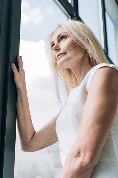 Bajo ángulo vista de elegante rubia madura mujer mirando a través de la ventana - foto de stock