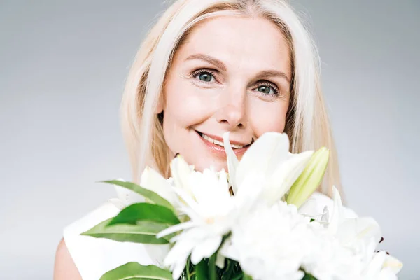 Mujer madura rubia feliz con flores blancas mirando a la cámara aislada en gris - foto de stock