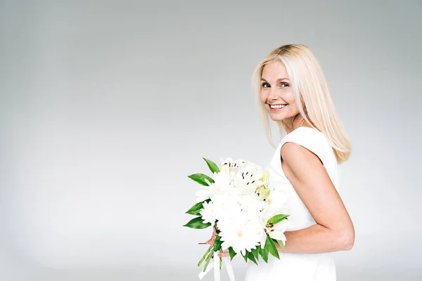 Side view of happy blonde mature woman with white bouquet isolated on grey — Stock Photo