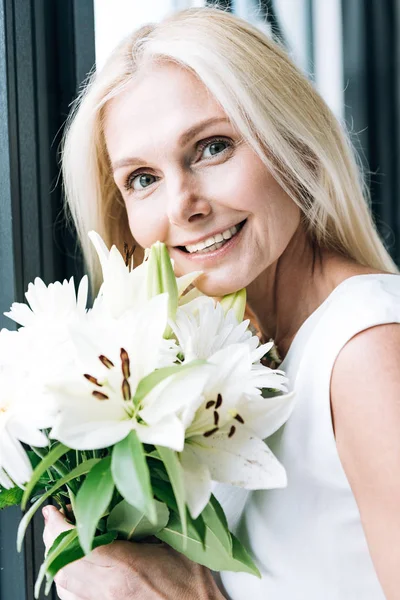 Porträt einer glücklichen blonden reifen Frau mit weißen Blumen am Fenster — Stockfoto