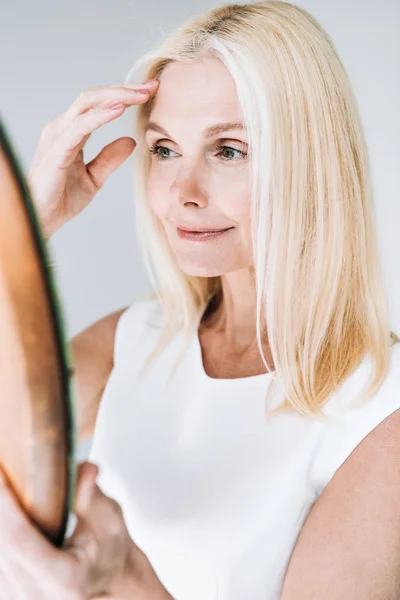 Happy blonde mature woman with mirror isolated on grey — Stock Photo