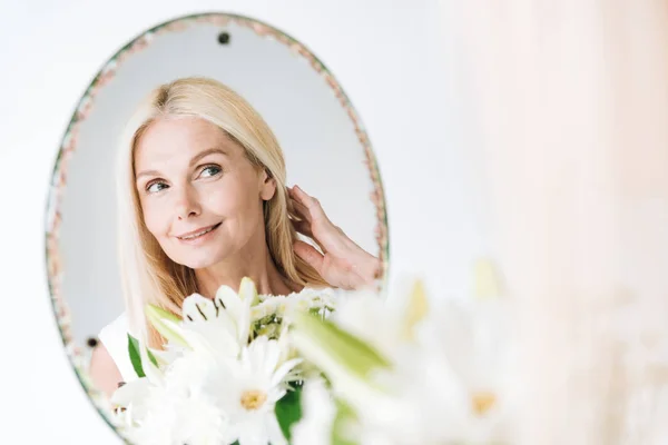 Happy blonde mature woman with flowers looking through mirror isolated on white — Stock Photo