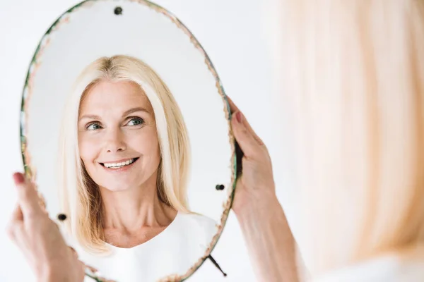 Heureux blonde mature femme regardant à travers miroir isolé sur blanc — Photo de stock