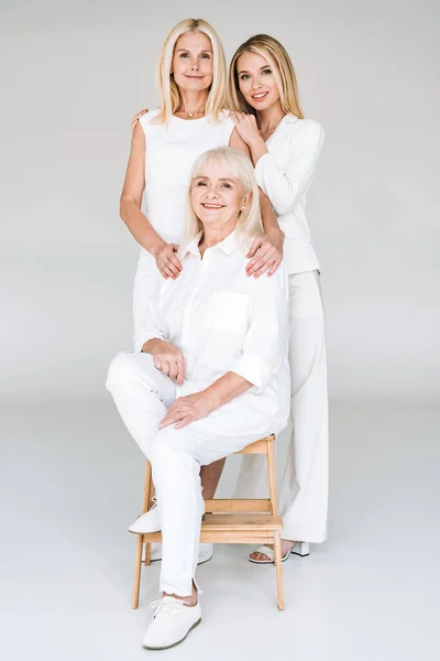 Rubia madre e hija de pie cerca de la abuela en silla de madera sobre fondo gris - foto de stock