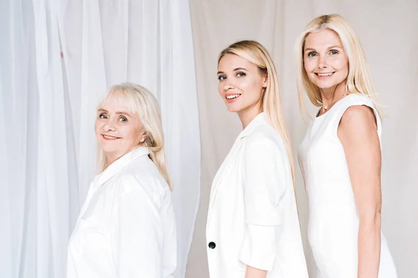 Vista lateral de la sonrisa elegante de tres generaciones de mujeres rubias en trajes blancos totales mirando a la cámara - foto de stock