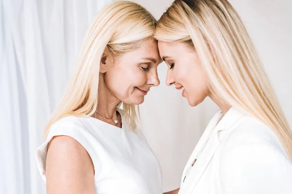 Sonriente elegante rubia madre e hija en total blanco trajes de pie cara a cara con los ojos cerrados - foto de stock