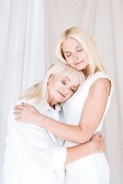 Erfreut elegante blonde reife Tochter und ältere Mutter in ganz weißen Outfits, die sich mit geschlossenen Augen umarmen — Stockfoto