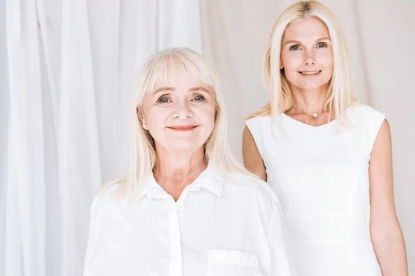 Smiling elegant blonde mature daughter and senior mother in total white outfits — Stock Photo