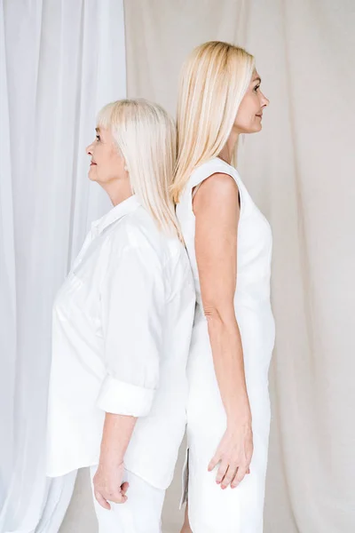 Side view of blonde mature daughter and senior mother in total white outfits standing back to back — Stock Photo