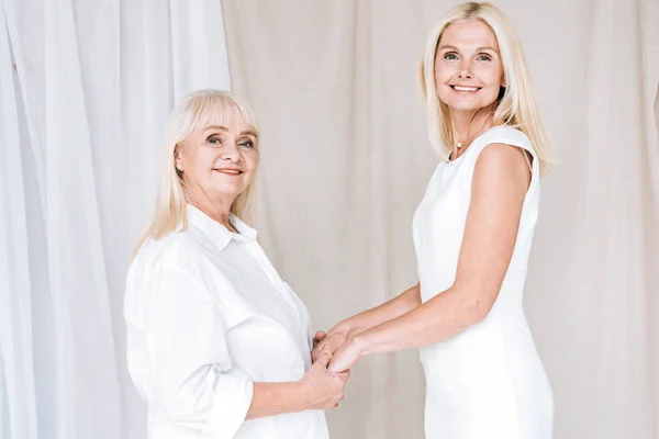 Feliz elegante rubia madura hija y senior madre en total blanco trajes cogidos de la mano - foto de stock