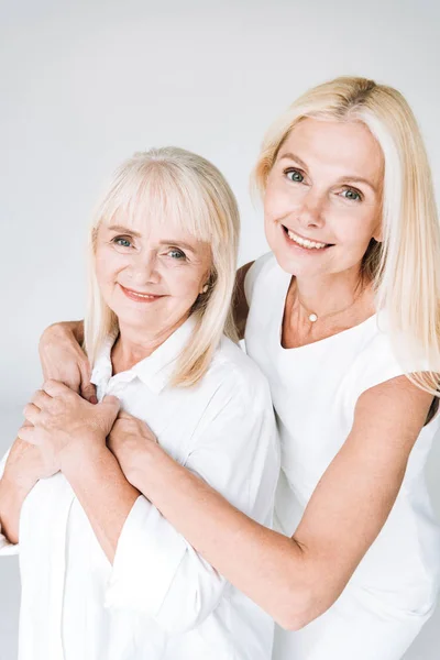 Elegante rubia madura hija y senior madre en total blanco trajes abrazando aislado en gris - foto de stock