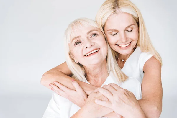 Feliz rubia madura hija y senior madre en total blanco trajes abrazando aislado en gris - foto de stock