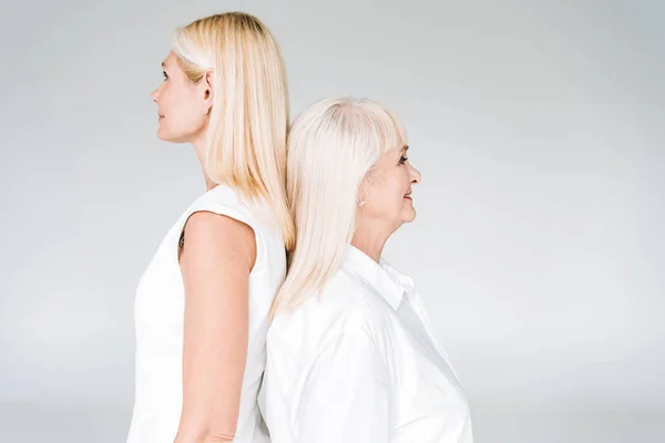 Elegante blonde reife Tochter und ältere Mutter in total weißen Outfits, die Rücken an Rücken isoliert auf grau stehen — Stockfoto