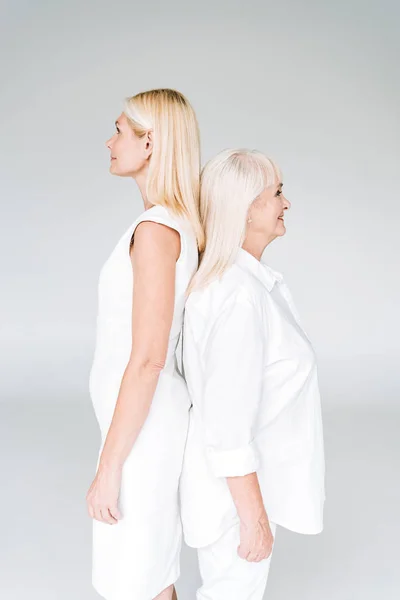 Blonde mature daughter and senior mother in total white outfits standing back to back isolated on grey — Stock Photo