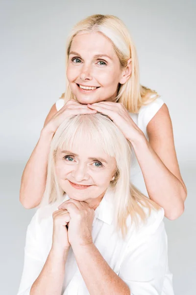 Улыбающаяся элегантная блондинка зрелая дочь и старшая мать в полностью белых нарядах, изолированных на сером — стоковое фото