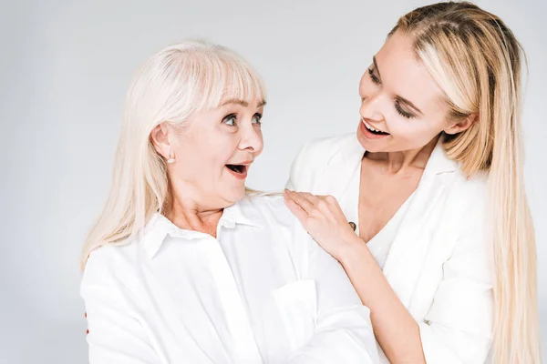 Збуджена блондинка бабуся і онука разом в загальному білому вбранні дивиться один на одного ізольовані на сірому — стокове фото