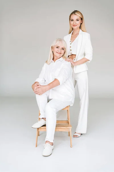 Vue pleine longueur de la grand-mère blonde heureuse assise sur la chaise près de la petite-fille dans des vêtements blancs totaux — Photo de stock