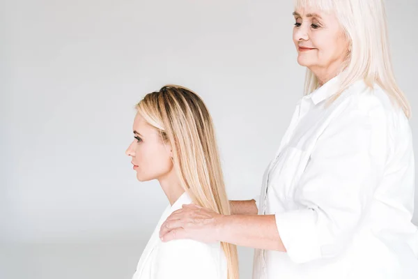 Side view of blonde grandmother putting hands on granddaughter shoulders isolated on grey — Stock Photo