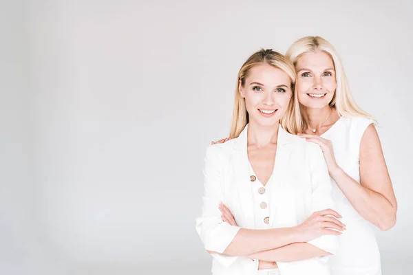 Smiling blonde mature mother embracing young daughter in total white clothes isolated on grey — Stock Photo