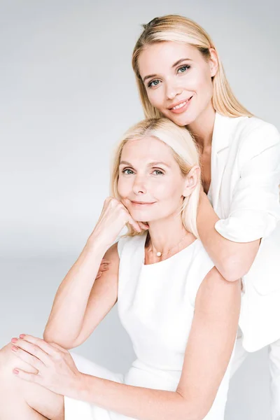 Smiling blonde young daughter embracing mature mother in total white clothes isolated on grey — Stock Photo